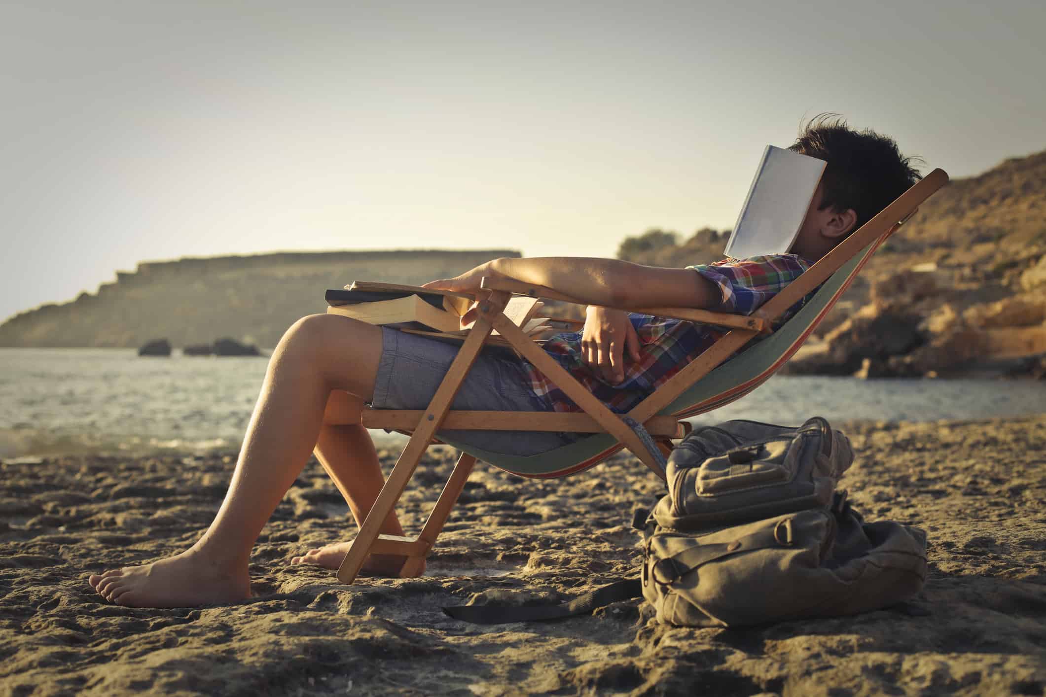 Young guy fell asleep while laying on the beach in a sunbathing chair, studying, having his face covered by his book and the others on his lap