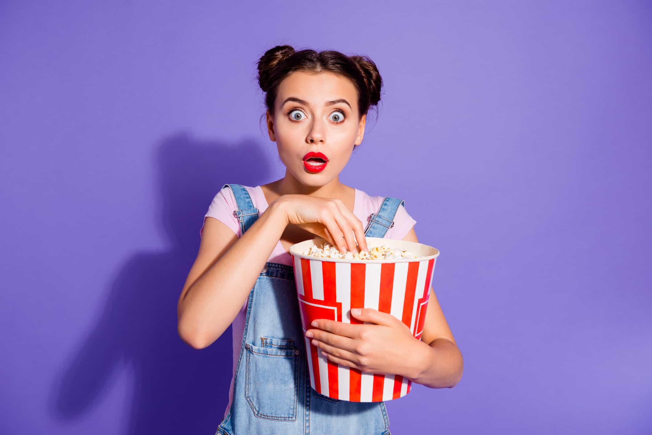 Close up photo beautiful amazing she her lady two buns watch tv show hold popcorn bucket eyes full fear mouth open wear casual t-shirt jeans denim overalls clothes isolated purple violet background